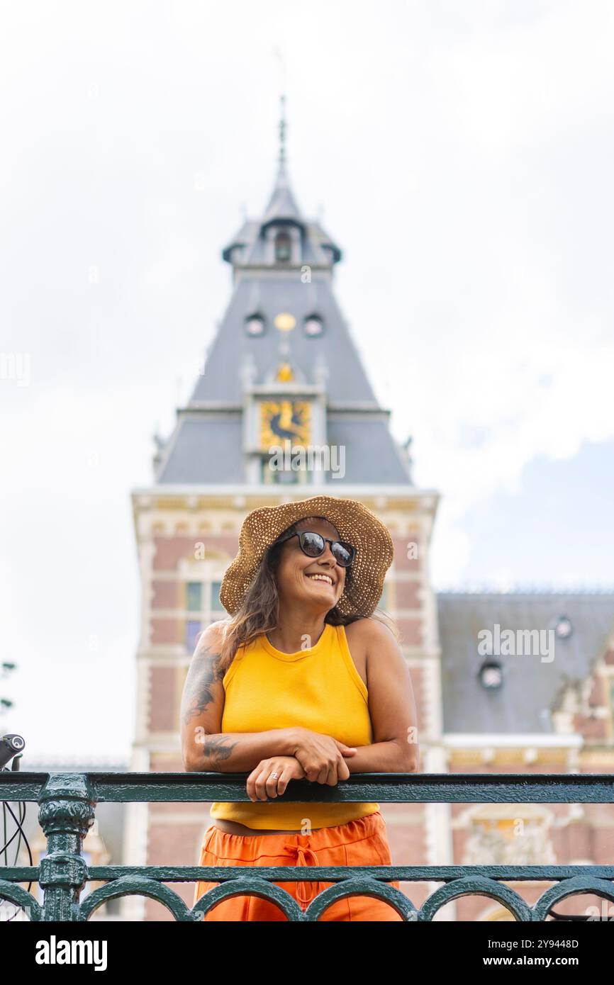 A cheerful woman in a straw hat and sunglasses smiles on a bridge with the iconic Amsterdam architecture in the background, representing a perfect fal Stock Photo