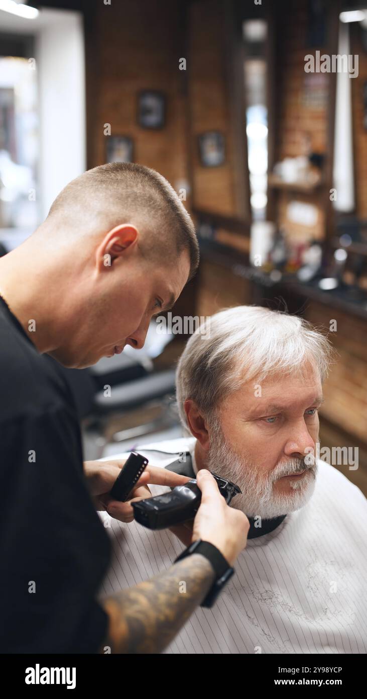 Young man, barber working with precision on client beard, using electric trimmer. Elderly man visiting barber shop Stock Photo