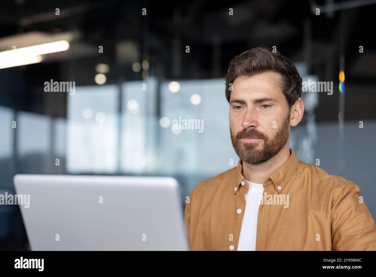 Man with beard using laptop in office. Reflects concentration, focus, productivity in modern workspace. Casual wear suggests informal but professional setting. Stock Photo