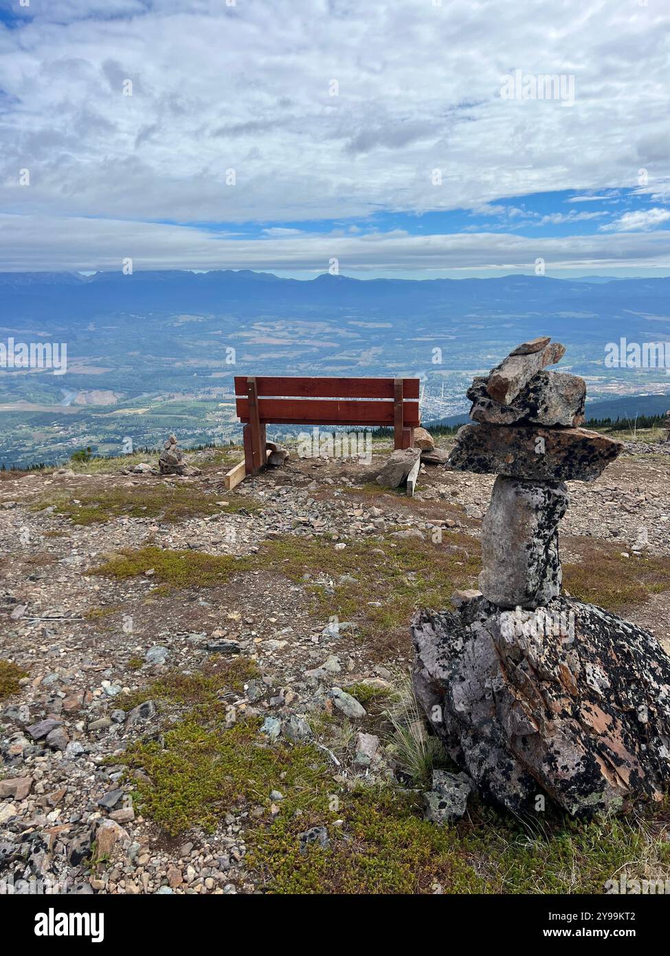 Wooden bench and Inukshuk overlooking a panoramic Smithers valley view from Hudson Bay Mountain, British Columbia, Canada, under cloudy, expansive sky - Smartphone Captured Stock Image