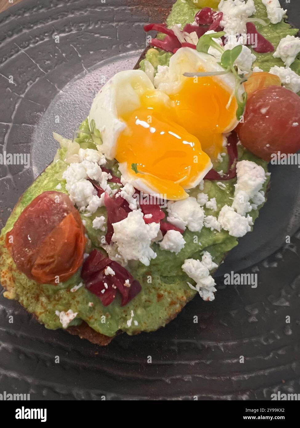 Avocado toast topped with a poached egg, feta cheese, roasted tomatoes, and microgreens, served on rustic bread for a fresh and delicious meal - Smartphone Captured Stock Image