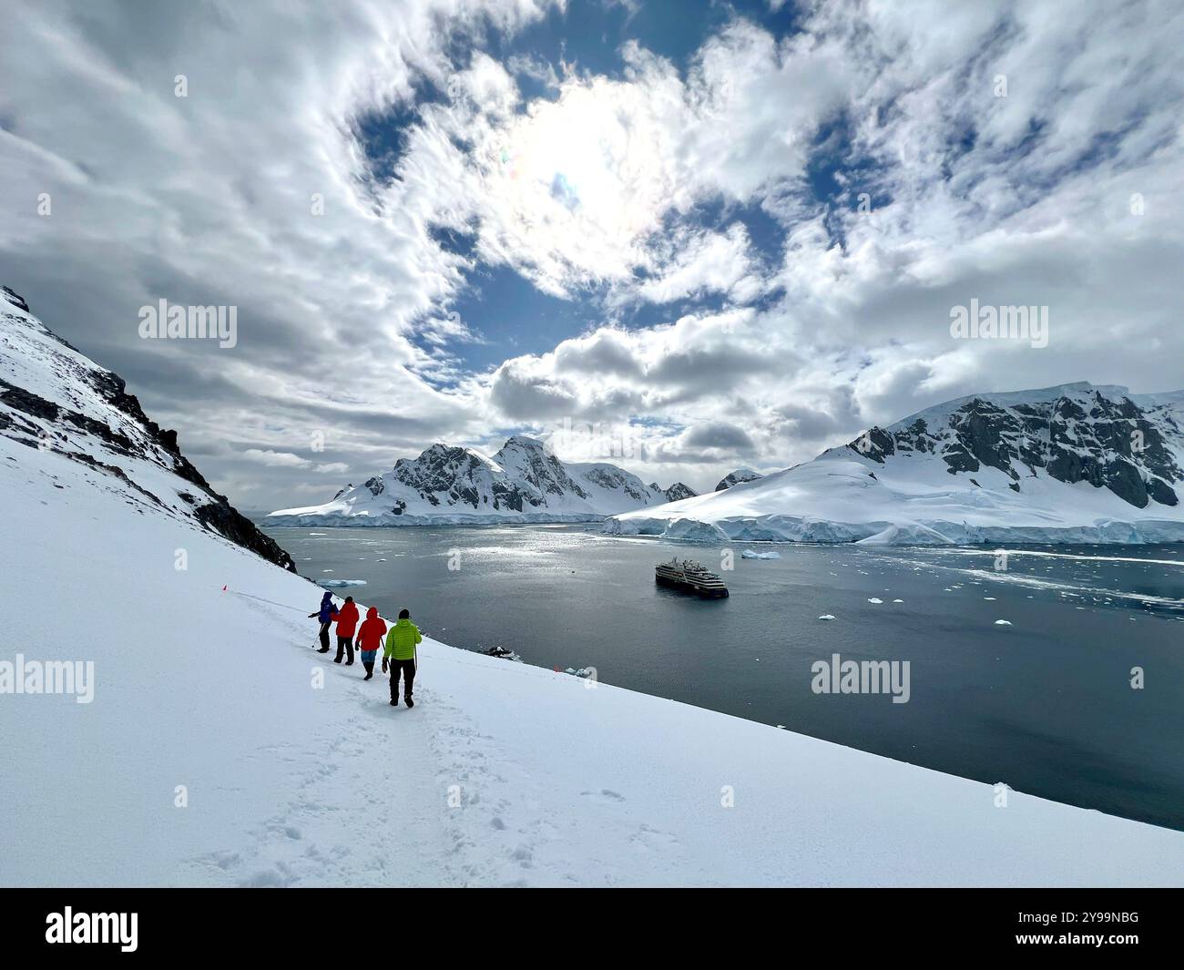 Hikers trek through snow-covered slopes overlooking National Geographic Endurance docked in a serene bay at Graham Land, Antarctica - Smartphone Captured Stock Image