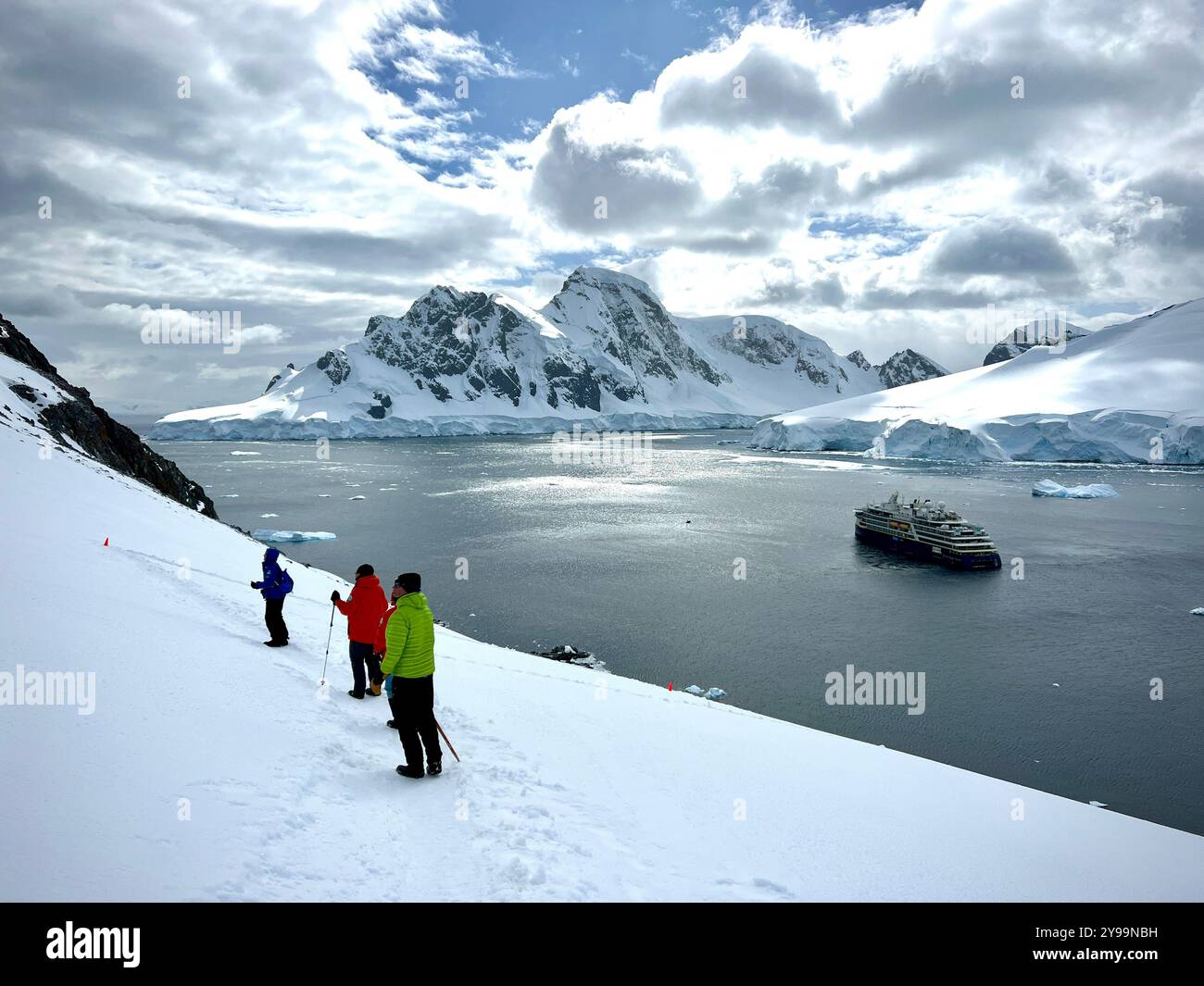 Hikers in vibrant gear descend snowy slopes overlooking National Geographic Endurance in a tranquil bay surrounded by towering glaciers, Graham Land - Smartphone Captured Stock Image