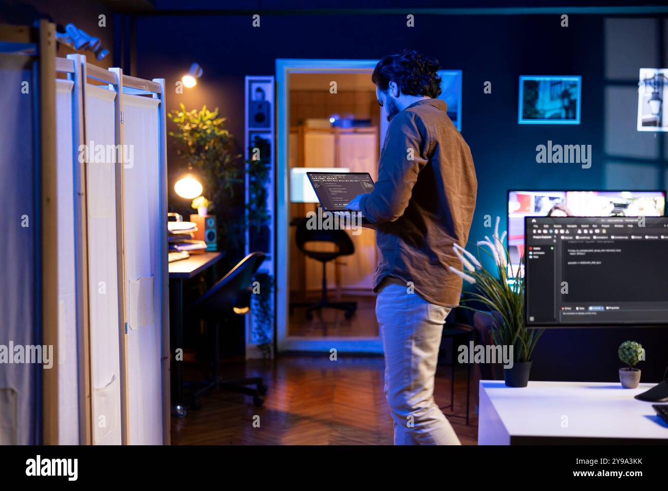 IT expert working from neon lit home using laptop, testing and deploying programs and systems. Developer checking code displayed on computer monitors, standing in apartment Stock Photo