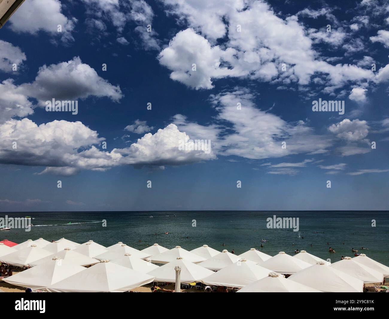 Rows of white umbrellas line the shore and virga on the background under a blue sky with fluffy clouds overlooking the Black Sea, Chornomorsk, Ukraine - Smartphone Captured Stock Image