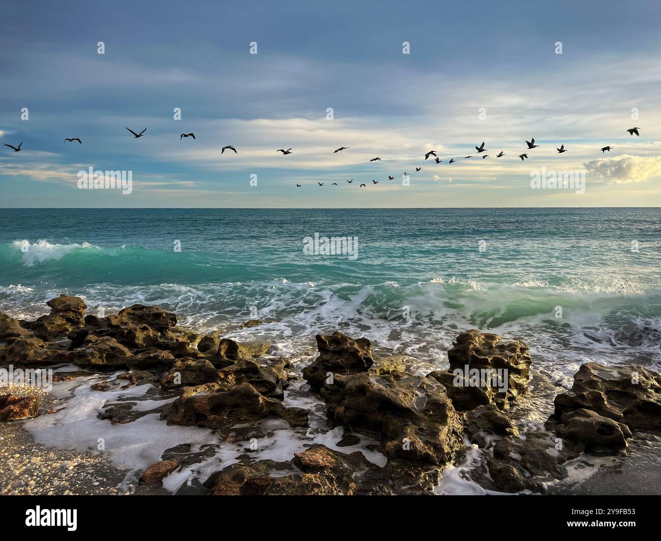 Jagged rocks line the foreground, partially embraced by the frothy turquoise waves of the ocean. Flocks of pelicans fly through this peaceful seascape - Smartphone Captured Stock Image