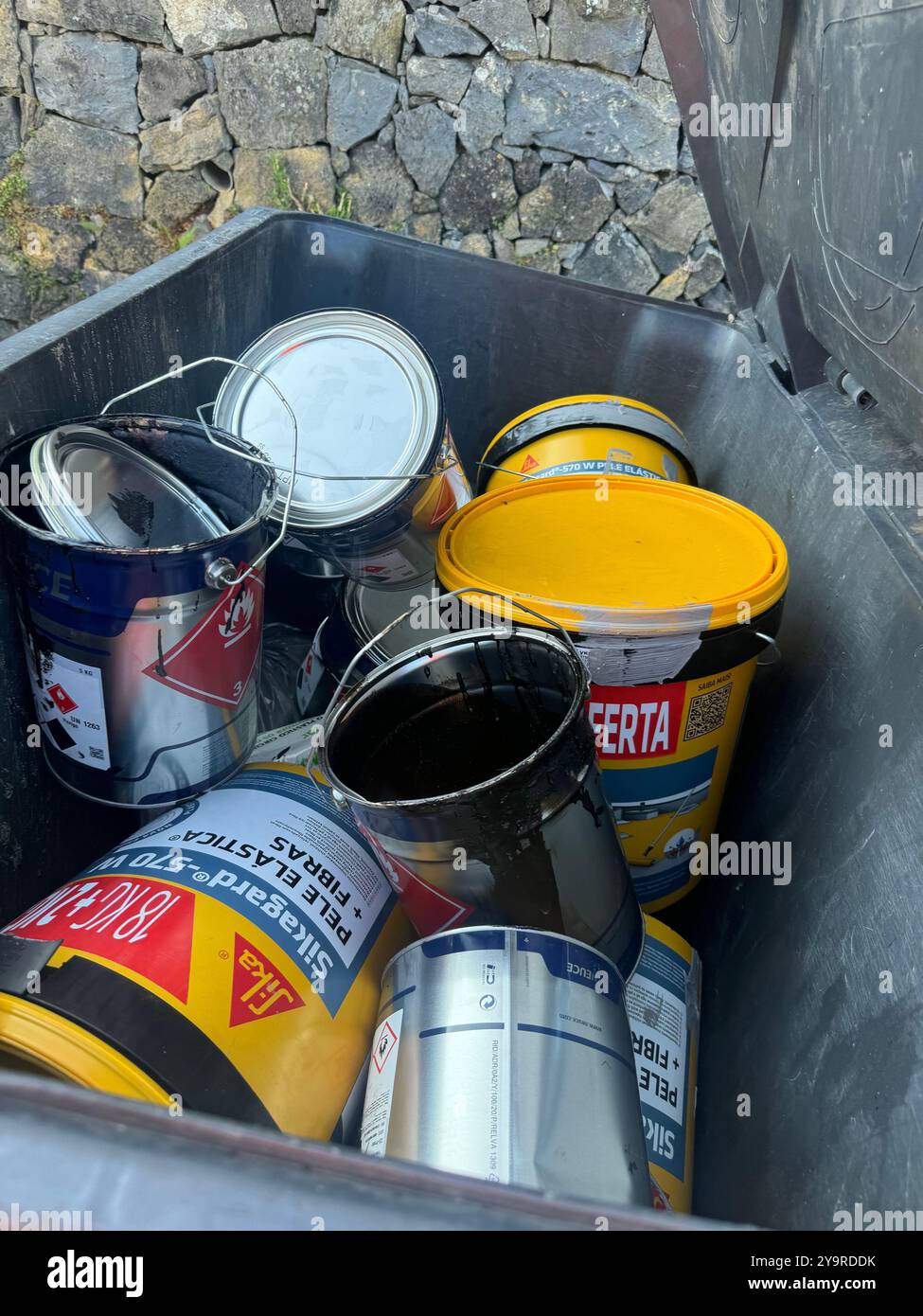 Discarded paint cans and buckets in an outdoor waste container against a stone wall. - Smartphone Captured Stock Image
