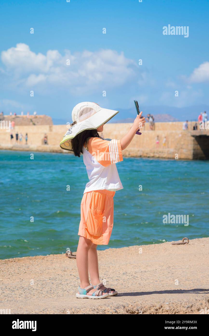 Child holiday, view of a young female child wearing a large summer hat taking a photo of the Inner Harbour area in Chania (Hania), Crete, Greece Stock Photo