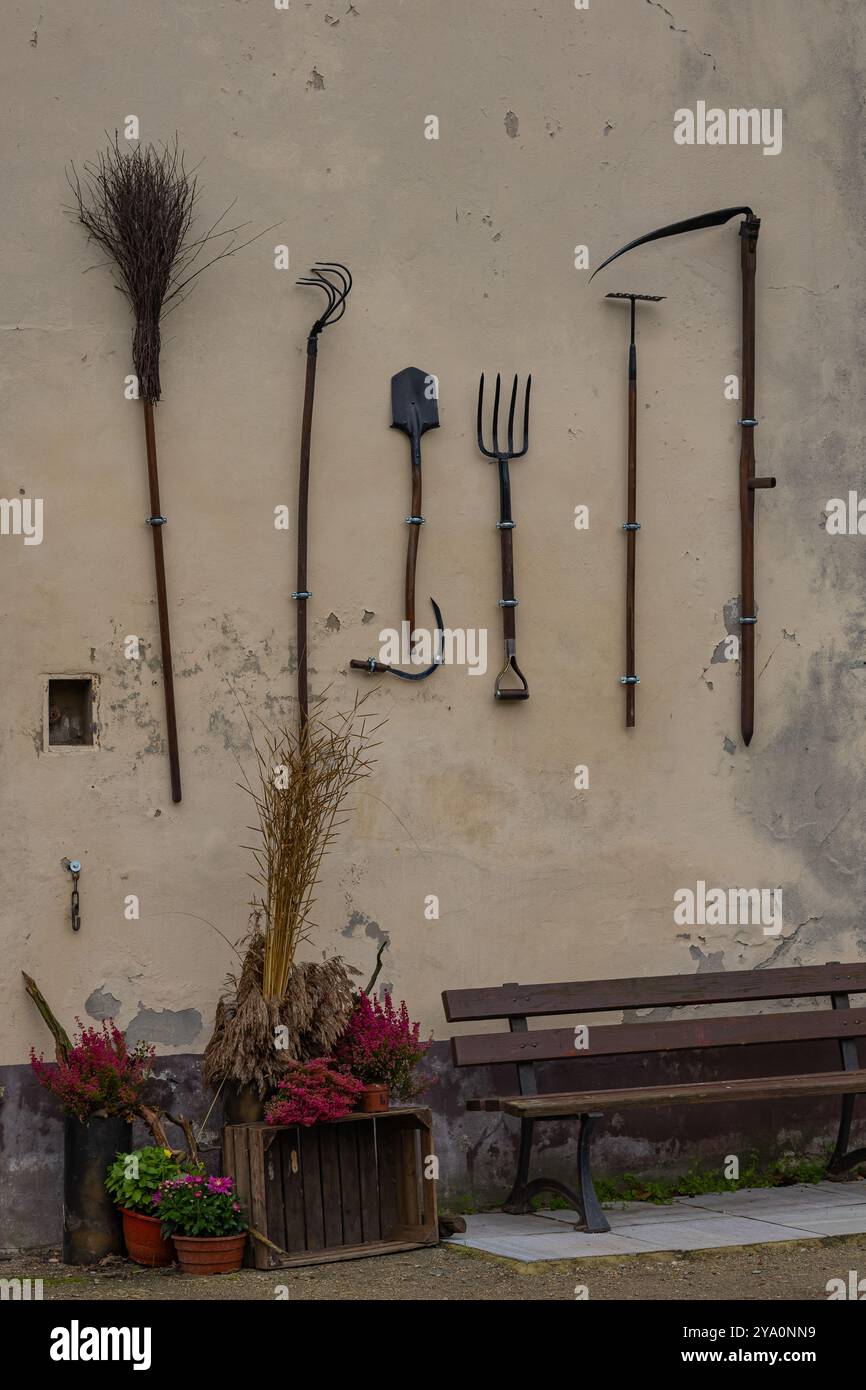 Vintage farming tools hanging on rustic wall with potted plants and a bench. Concept of traditional agriculture, rural lifestyle and craftsmanship Stock Photo