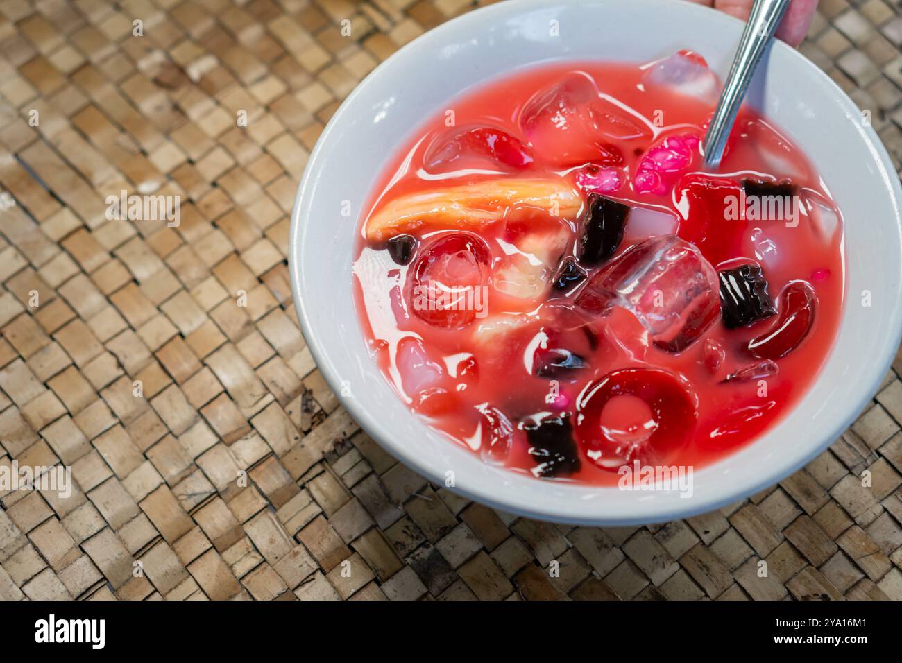 A bowl of colorful dessert featuring jelly pieces, fruits, and ice in a sweet syrup. The bowl is placed on a woven surface, highlighting the vibrant c Stock Photo