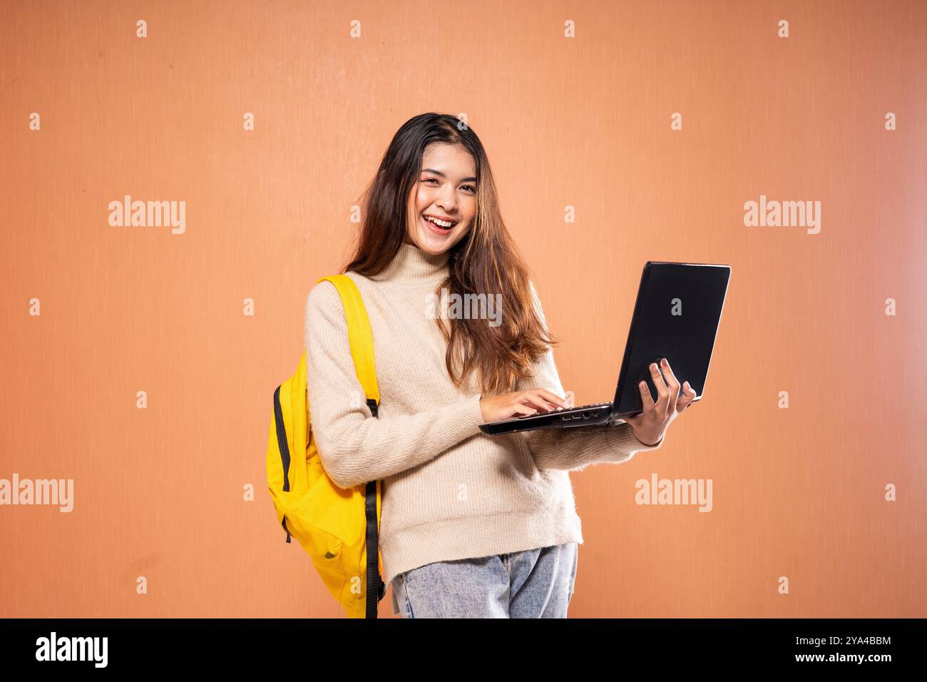 Cheerful Student Using Laptop with Yellow Backpack Against Orange Wall Stock Photo