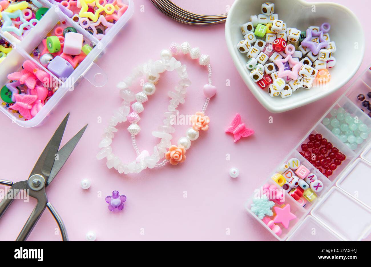 A creative arrangement of colorful beads, charms, and scissors on a pastel pink surface showcases the process of making unique jewelry. Delicate flora Stock Photo