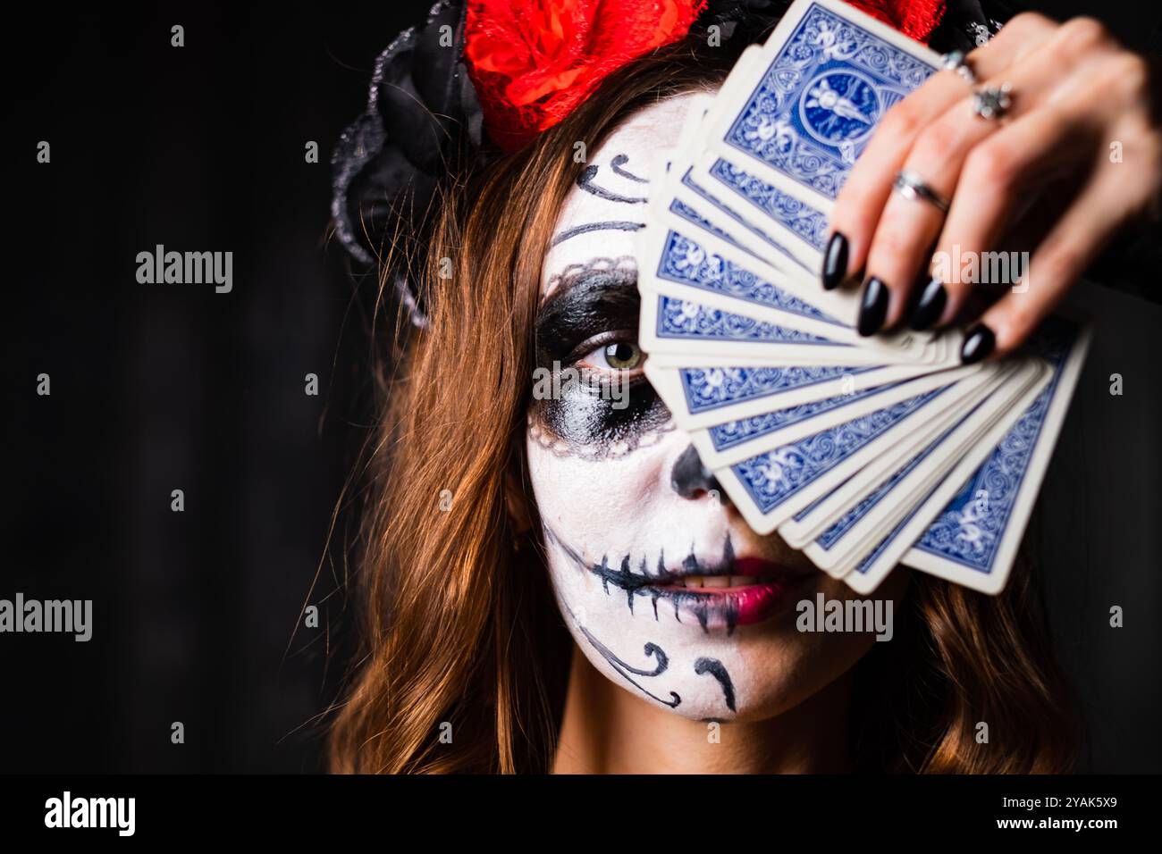 Woman is covering part of her face with playing cards, showing off her dia de los muertos sugar skull makeup. She has dark red hair and is wearing a black veil and red flower crown Stock Photo