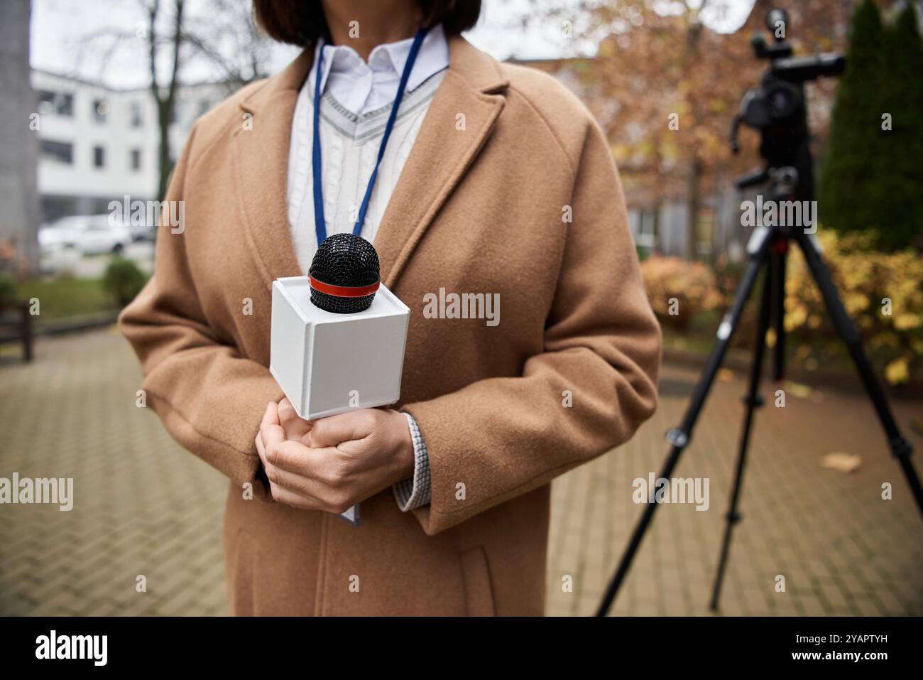 A focused journalist holding a microphone stands outdoor, preparing to deliver a news report amid autumn foliage. Stock Photo