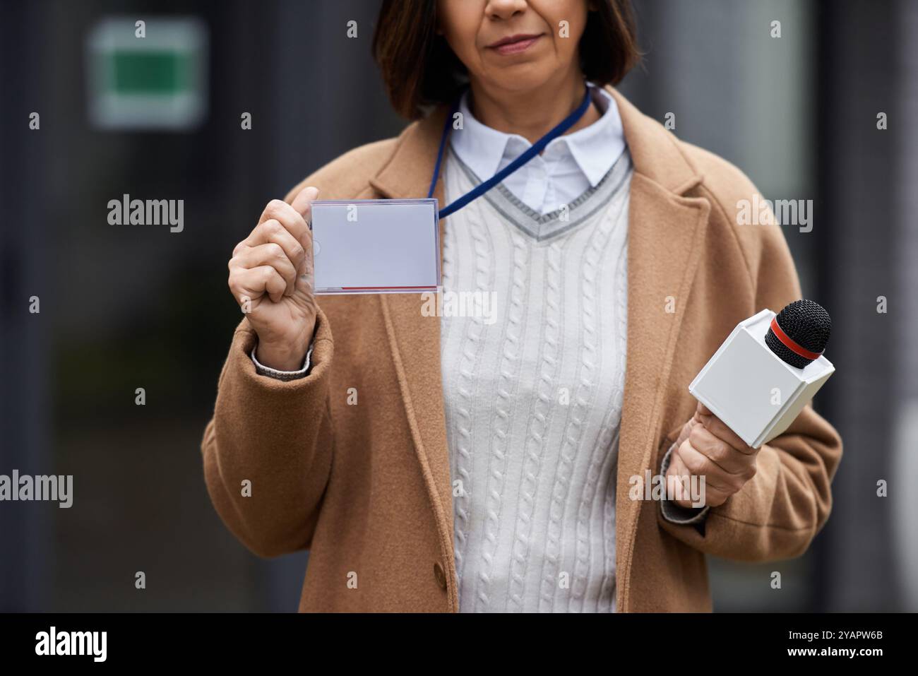 A multiracial female journalist holds a microphone while preparing to deliver a news report in the city, displaying her press badge Stock Photo