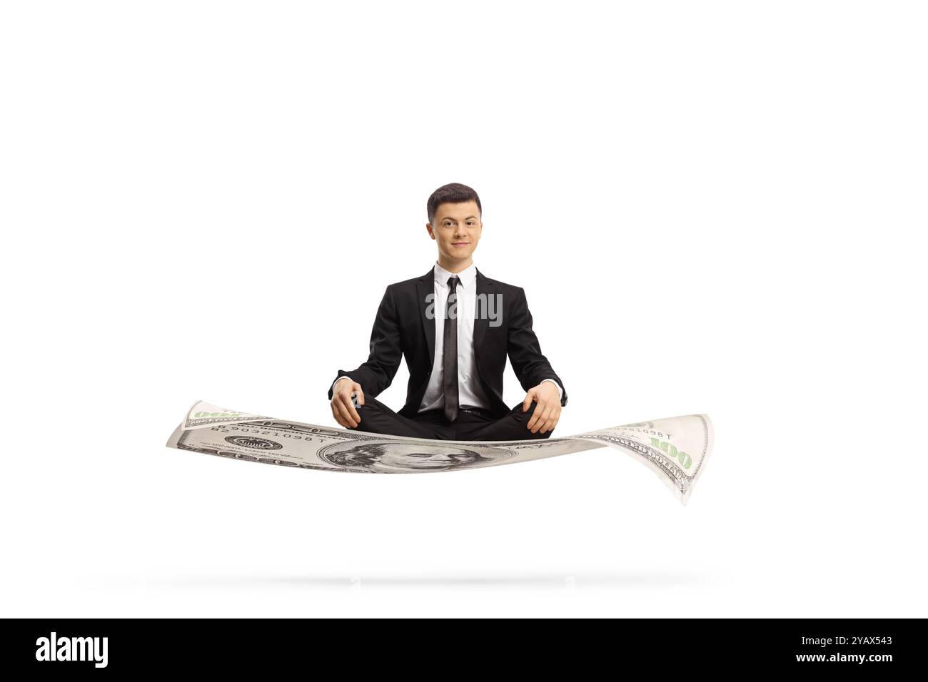 Young man in a black suit sitting on money with crossed legs isolated on white background Stock Photo