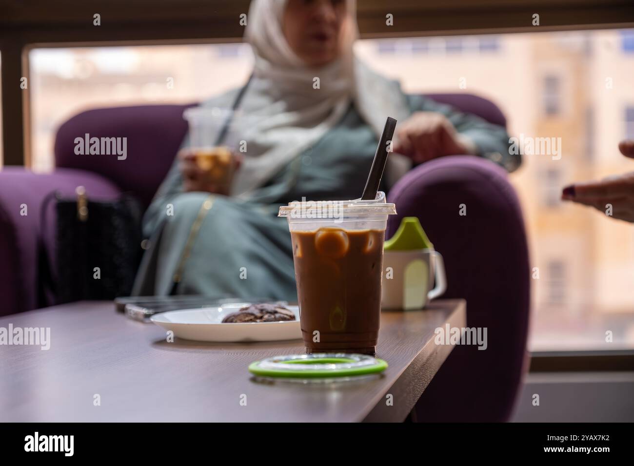 Chilled iced coffee served in a plastic cup with a black straw, accompanied by a chocolate cookie on a white plate. Perfect for a cozy coffee break Stock Photo