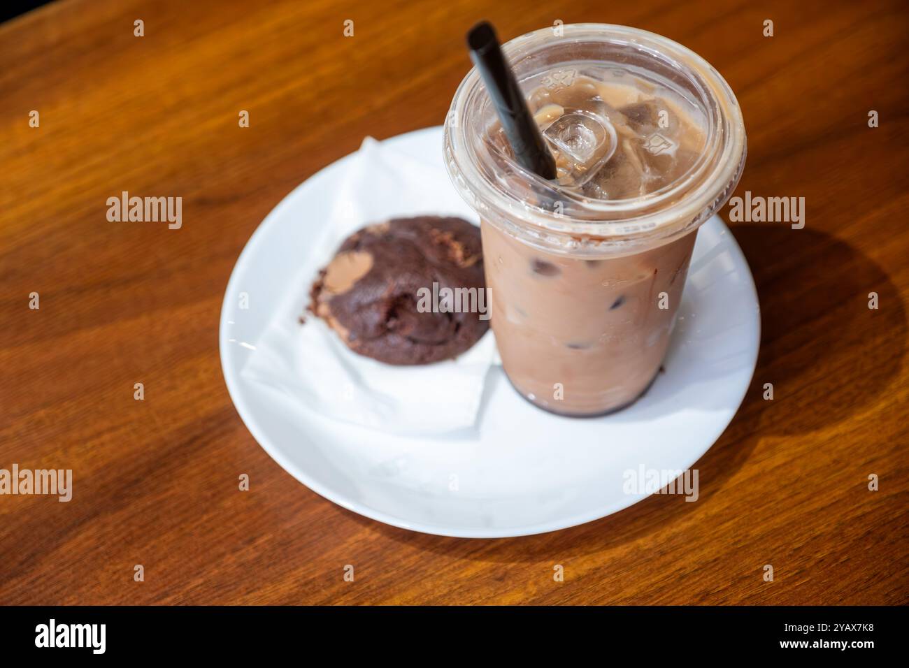 Chilled iced coffee served in a plastic cup with a black straw, accompanied by a chocolate cookie on a white plate. Perfect for a cozy coffee break Stock Photo