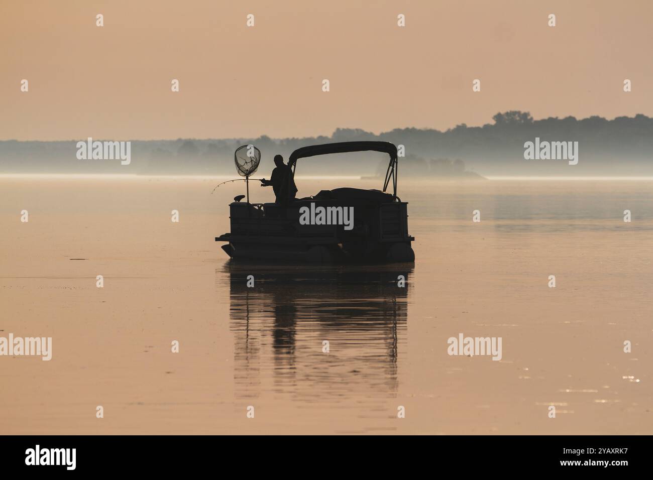 Silhouette of a man reeling in a fish on a pontoon boat during the early morning mist. Stock Photo