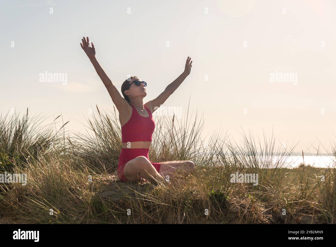 Woman sitting at the beach in the sand dunes with her arms outstretched enjoying the sun. Stress free concept. Stock Photo