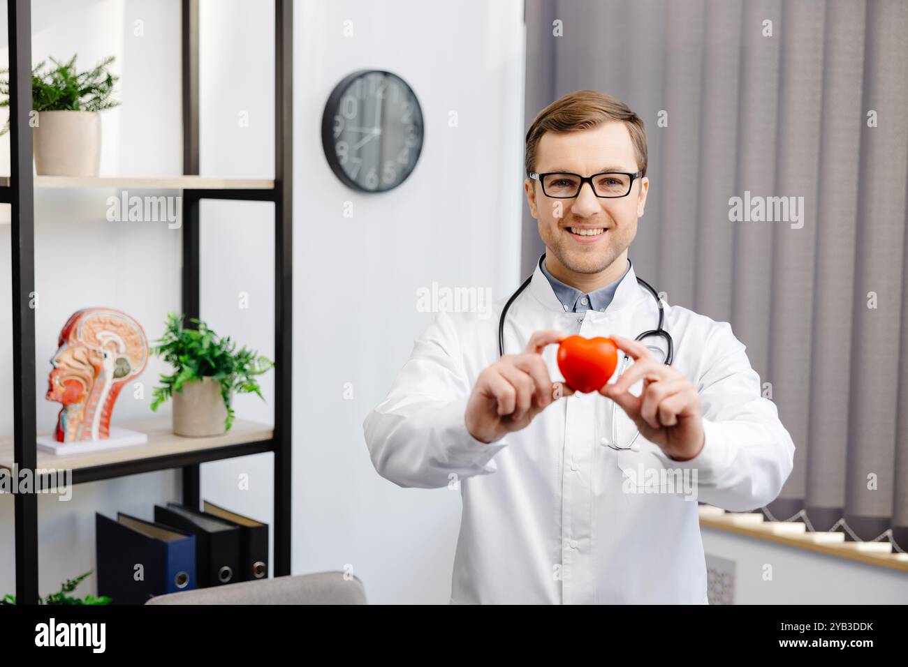 Doctor in white coat and glasses holding a red heart at hospital office ...
