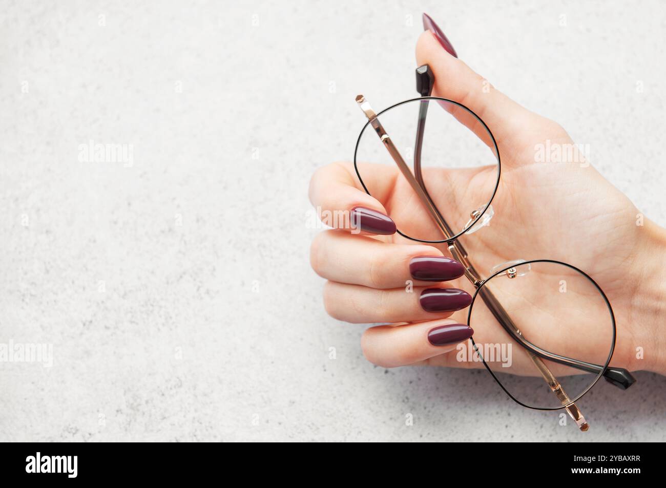 A hand with long burgundy nails gracefully holds a pair of round glasses above a light textured surface, illuminated by soft natural light. Stock Photo