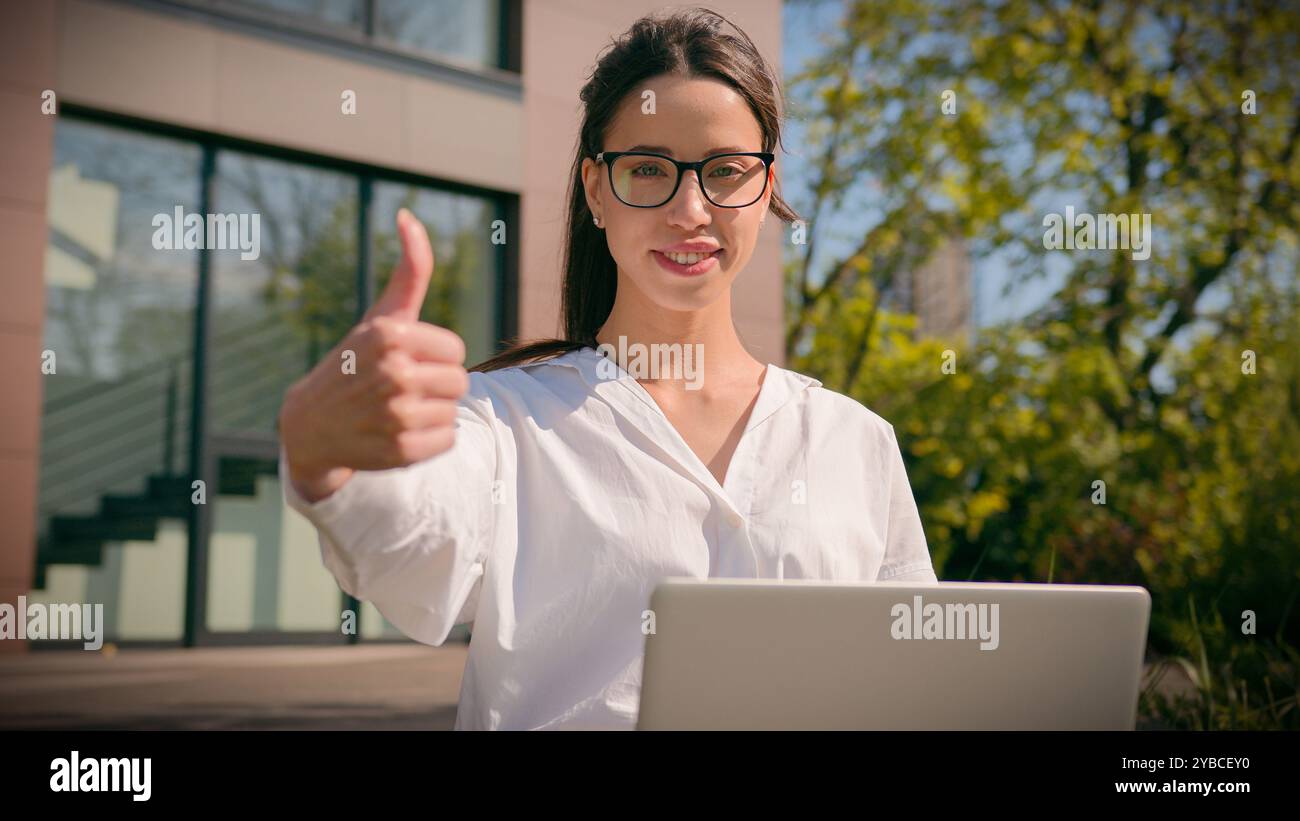 Smiling Caucasian woman girl female businesswoman freelancer student using laptop in city outdoors showing thumbs up gesture approval recommending Stock Photo