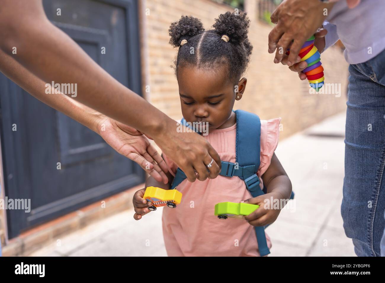 Baby african girl with schoolbag holding car toys standing in the street Stock Photo