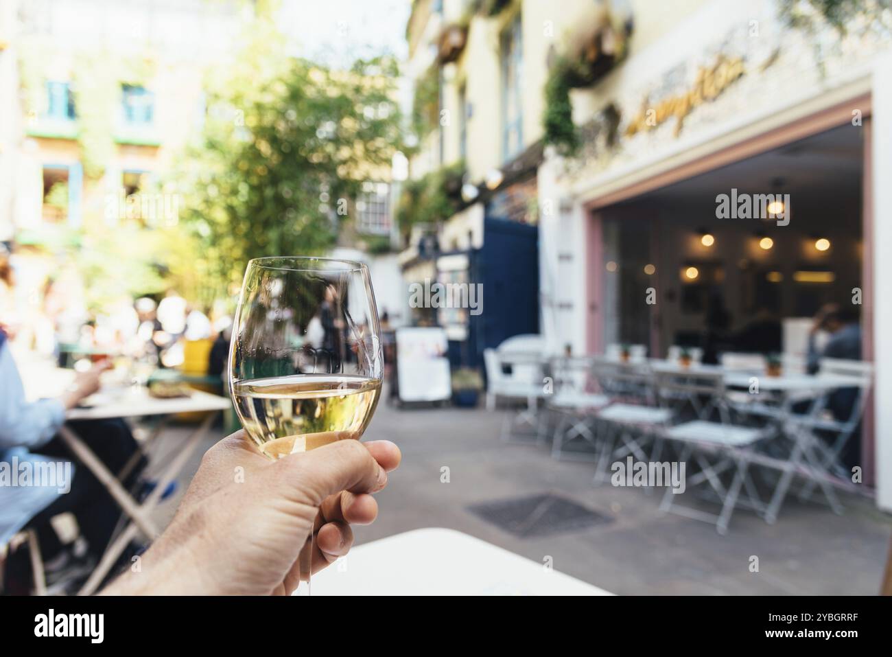 Man holds a glass of white wine in a trendy shopping area sitting on a terrace of a sidewalk cafe Stock Photo