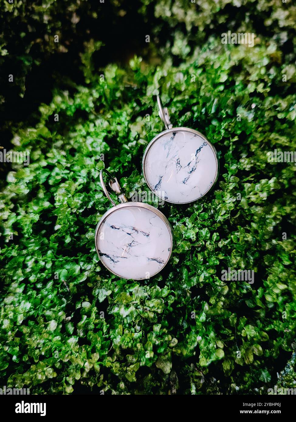 Handmade resin round marble texture stone earrings transparent finish at green moss background close. Leverback hooks, nature-inspired jewelry - Smartphone Captured Stock Image