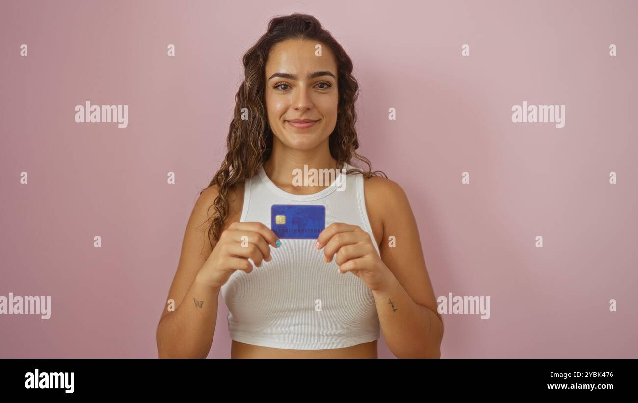 Young woman holding credit card in front of isolated pink background, smiling confidently and wearing white crop top Stock Photo
