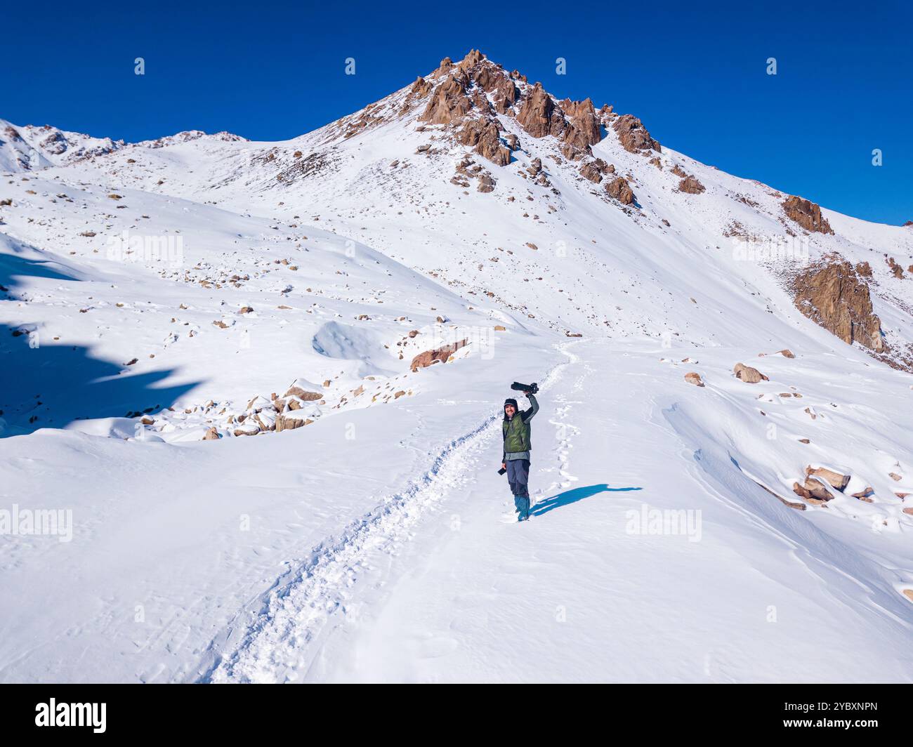 Joyful guy with camera raised above his head high in the mountains on a sunny day. Extreme pastime in snowy winter mountains Stock Photo