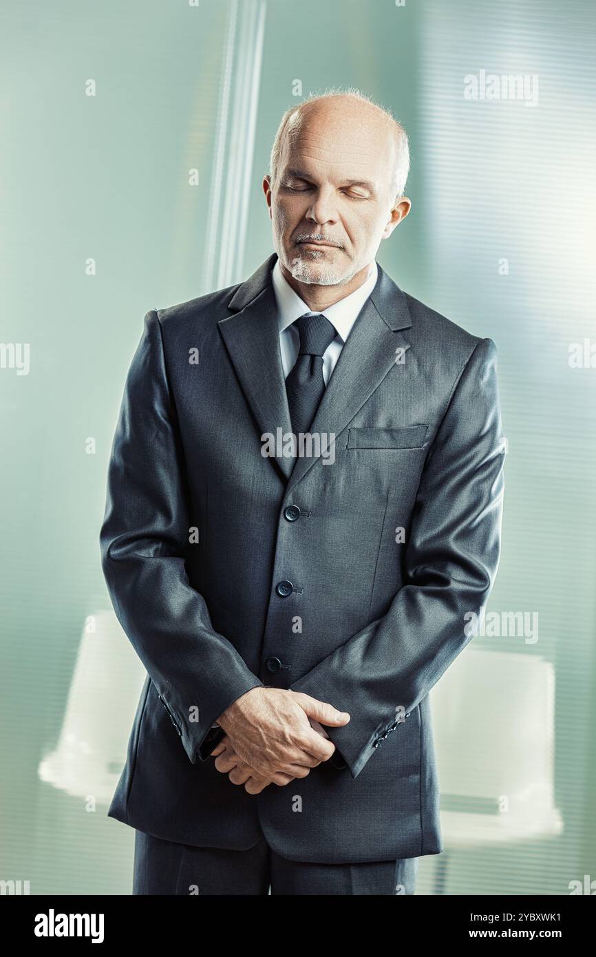 Senior businessman standing in a modern office, eyes closed, reflecting calmly. The scene exudes mindfulness and focus, showing confidence and leaders Stock Photo