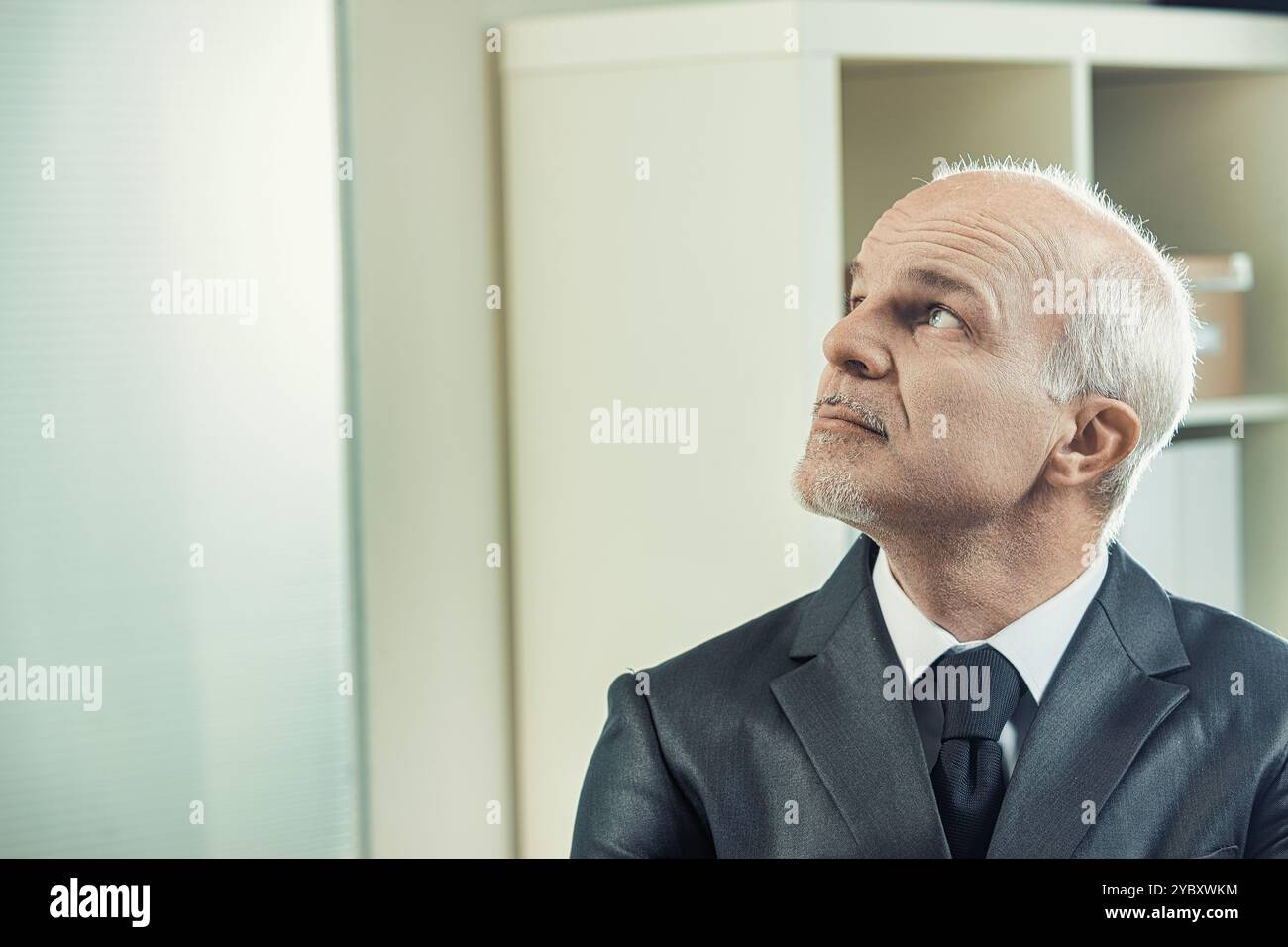 Senior businessman in a suit looks up thoughtfully in an office setting ...