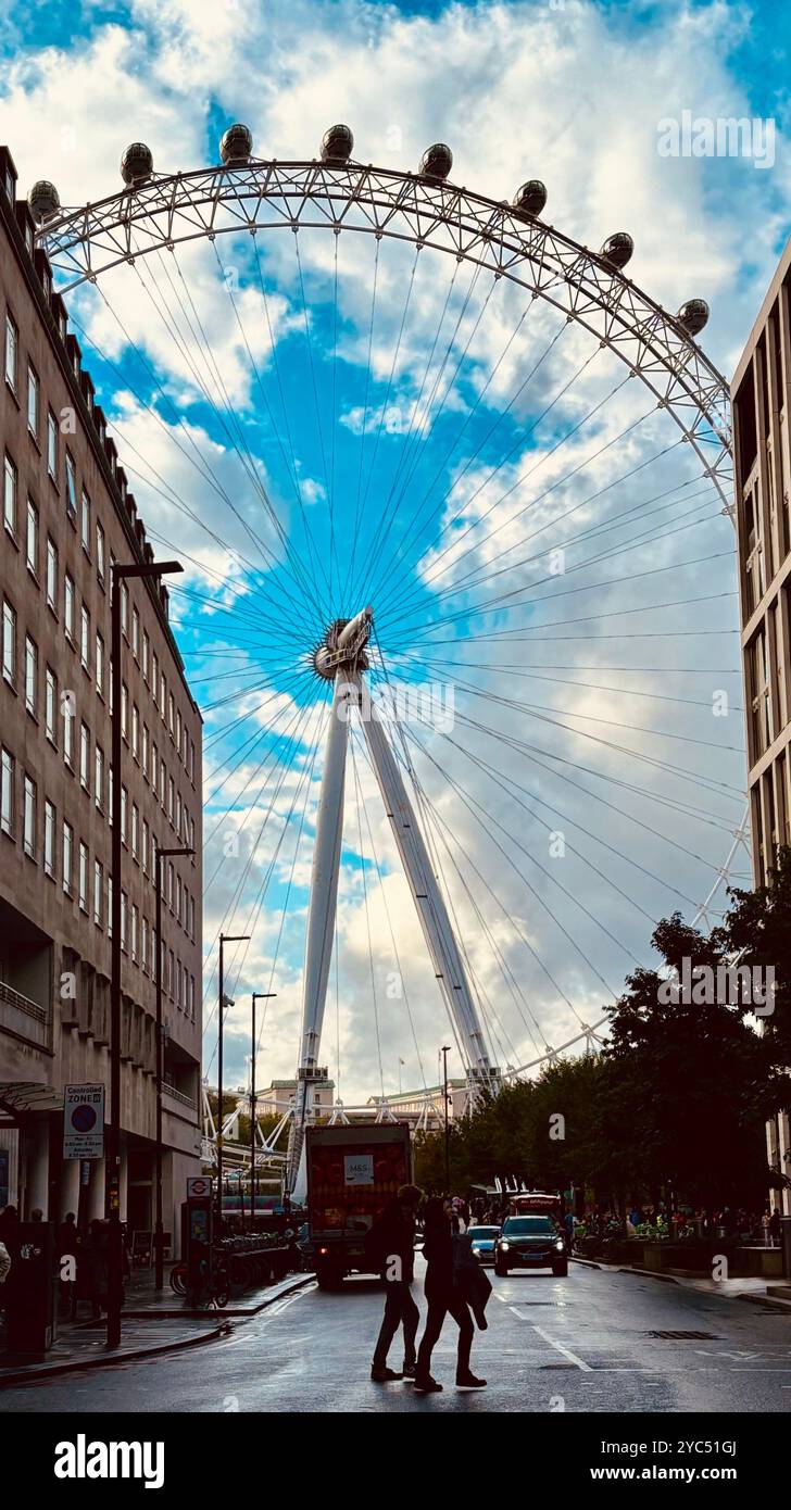A scenic view of the London Eye framed between city buildings with a cloudy sky, showcasing urban life and a busy street with pedestrians and vehicles - Smartphone Captured Stock Image A scenic view of the London Eye framed between city buildings with a cloudy sky, showcasing urban life and a busy street with pedestrians and vehicles - Smartphone Captured Stock Image