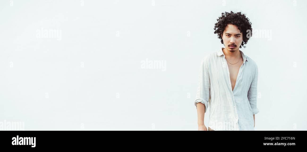 Young asian man with curly hair wearing a light, unbuttoned linen shirt stands confidently against a clean, white background. Ample copy space area to Stock Photo