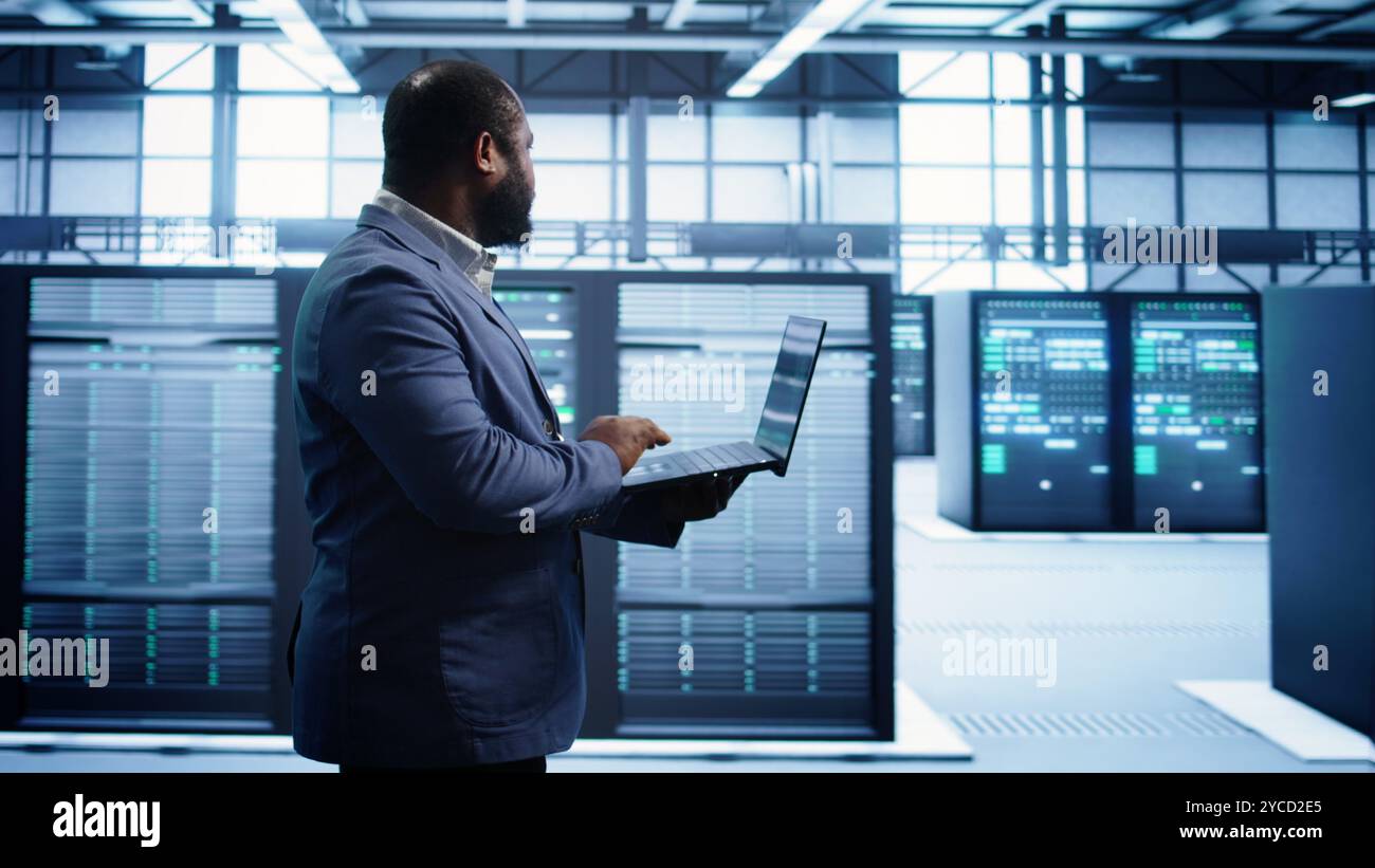 African american man walking in high tech facility, using laptop to patch network issues leading to slow data transfer rates. Data center worker fixing errors affecting supercomputers, camera B Stock Photo