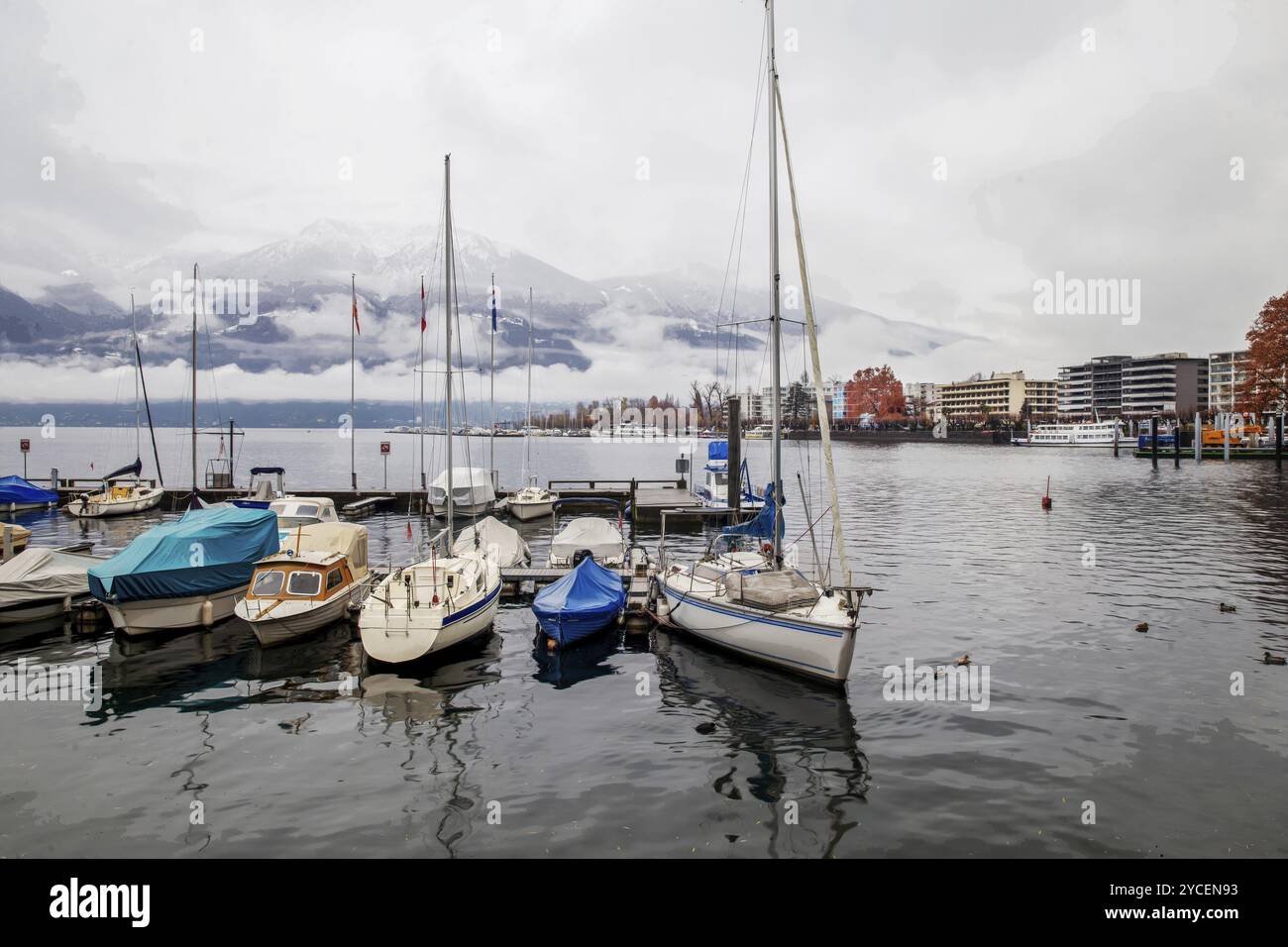 Fogging nature scene and sheltered sailboat in small harbor and pier in winter water and orange larch trees in distance and town Locarno Stock Photo