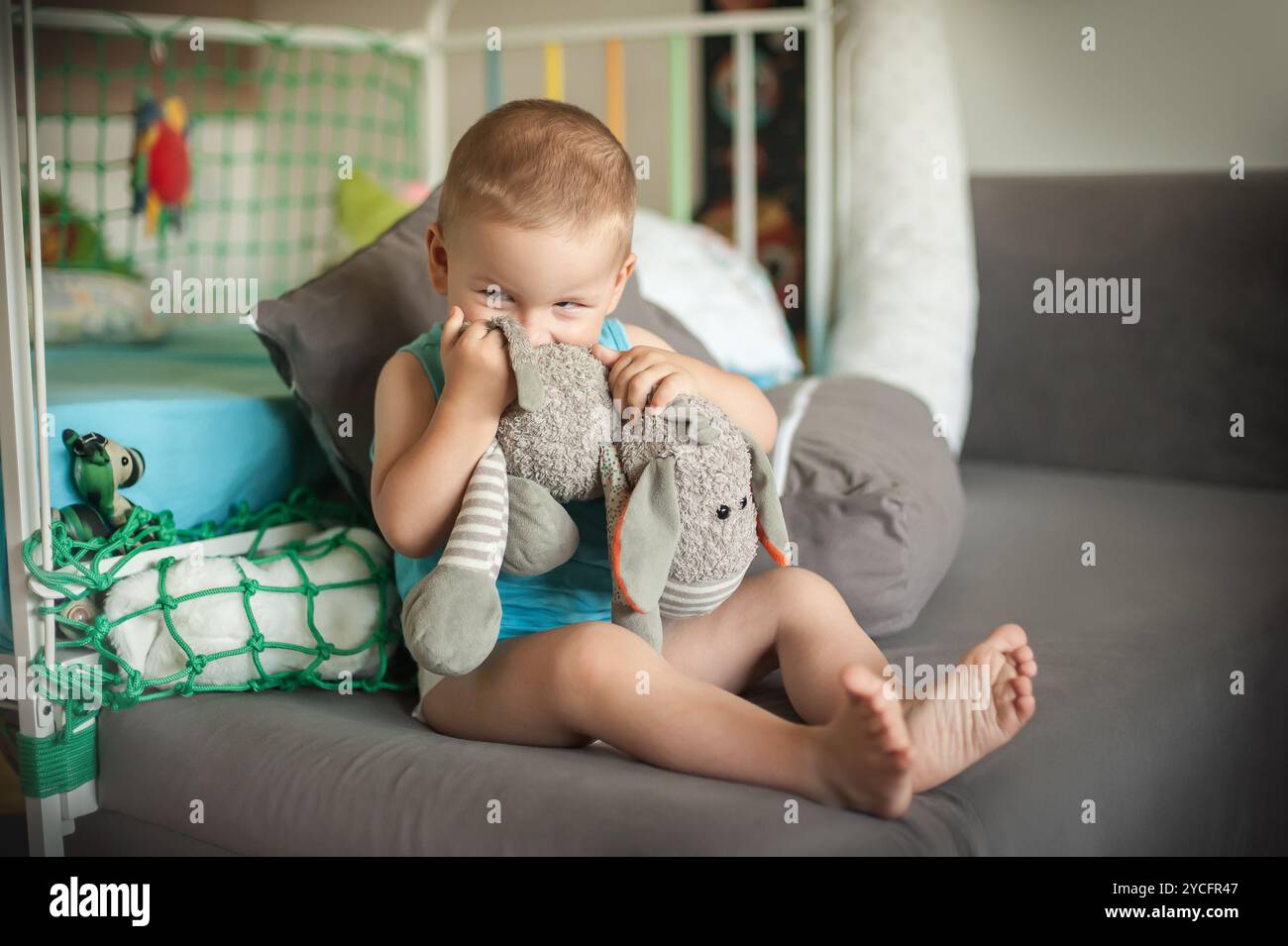 The cheerful toddler sits on a couch, hugging a gray stuffed elephant tightly while smiling, creating a warm atmosphere in the bright room. Stock Photo