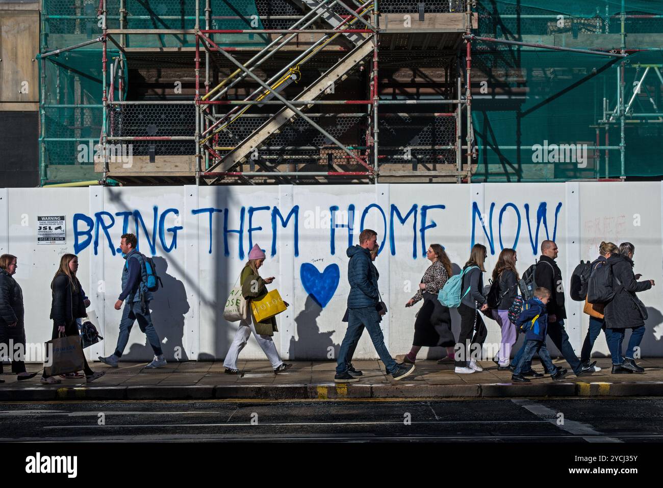 Pedestrians walking by "Bring Them Home Now" graffiti on boards in front of the Jenners redevelopment on Princes Street, Edinburgh, Scotland, UK. Stock Photo