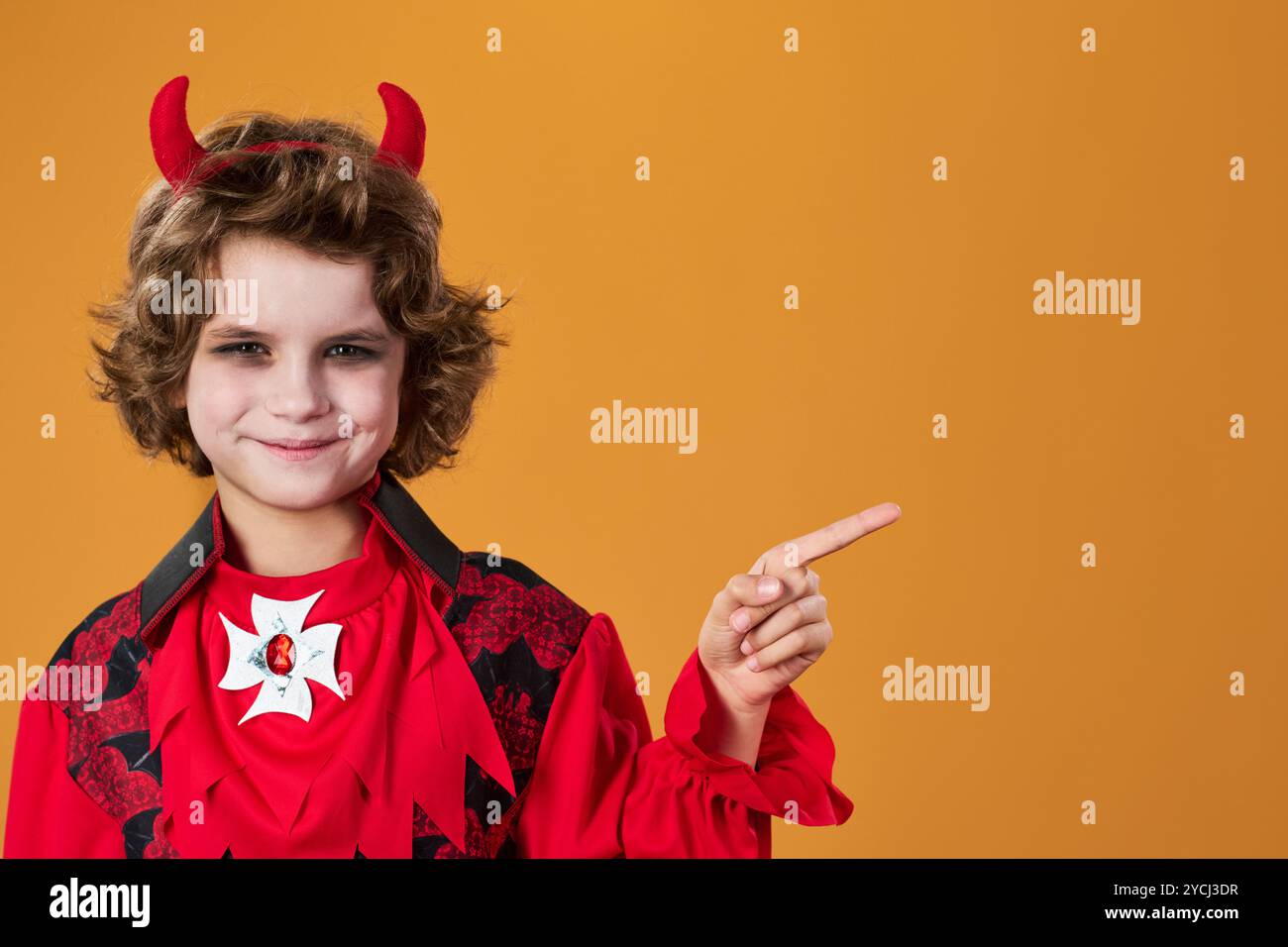 Youthful smiling boy with red horns of Halloween vampire or demon pointing aside at copyspace for your advert or offer Stock Photo