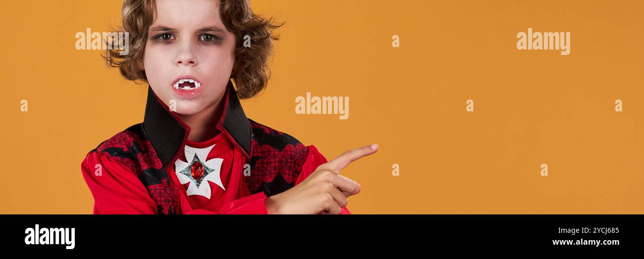 Header of scary youthful demon or vampire with red horns looking at you and pointing aside at copyspace over orange background Stock Photo