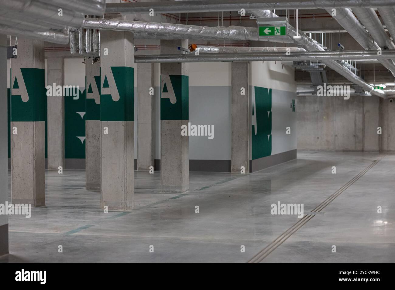 Empty underground parking garage with concrete pillars and ventilation pipes, marked with green letters and emergency exit signs Stock Photo