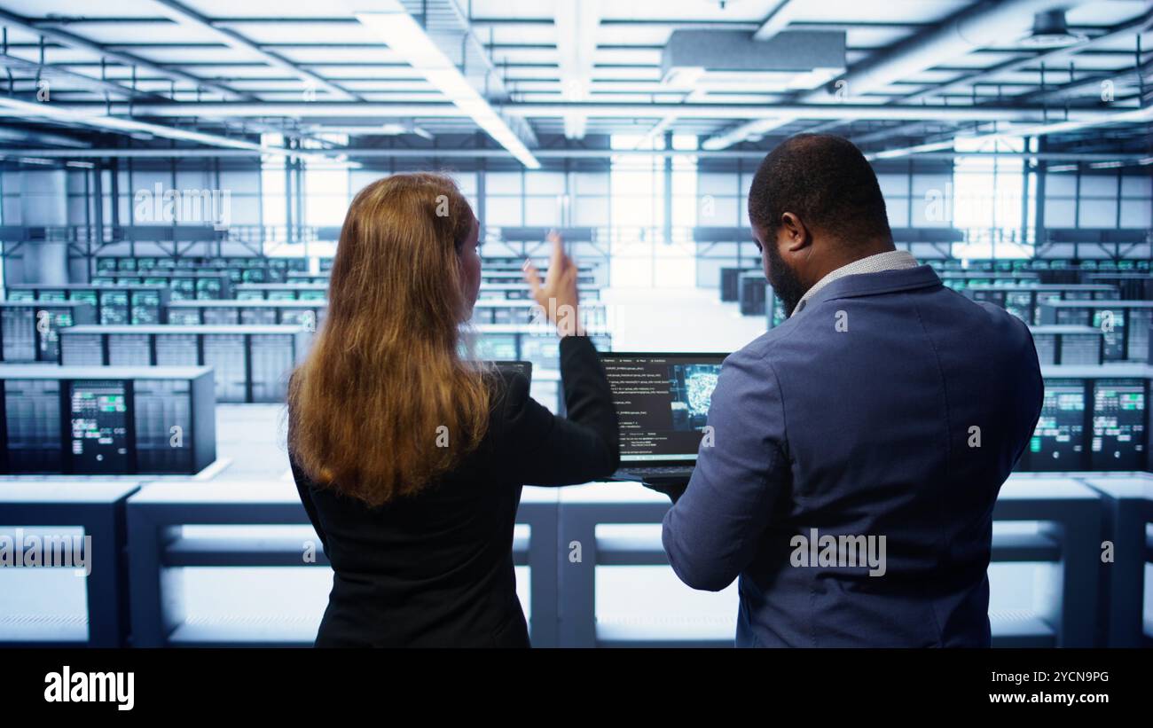 Teamworking programmers in data center overseeing server operations using laptop and tablet. Coworkers ensuring optimal performance and data integrity in server room using devices, camera B Stock Photo