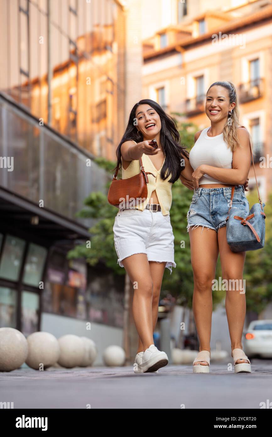 Two young women are walking down a city street, one of them is pointing at something and they are both smiling Stock Photo