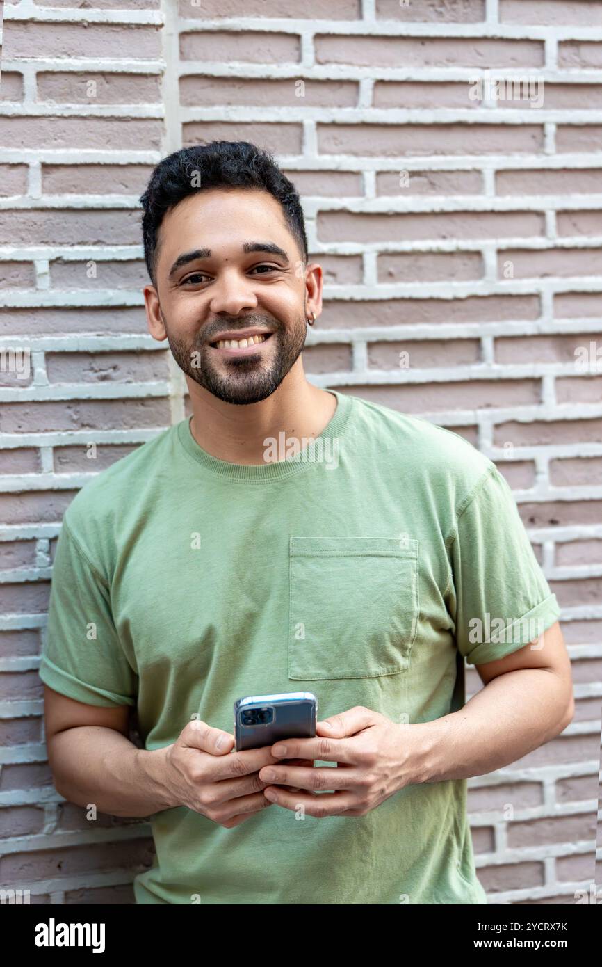 Young man is smiling while using a smartphone and leaning against a brick wall Stock Photo