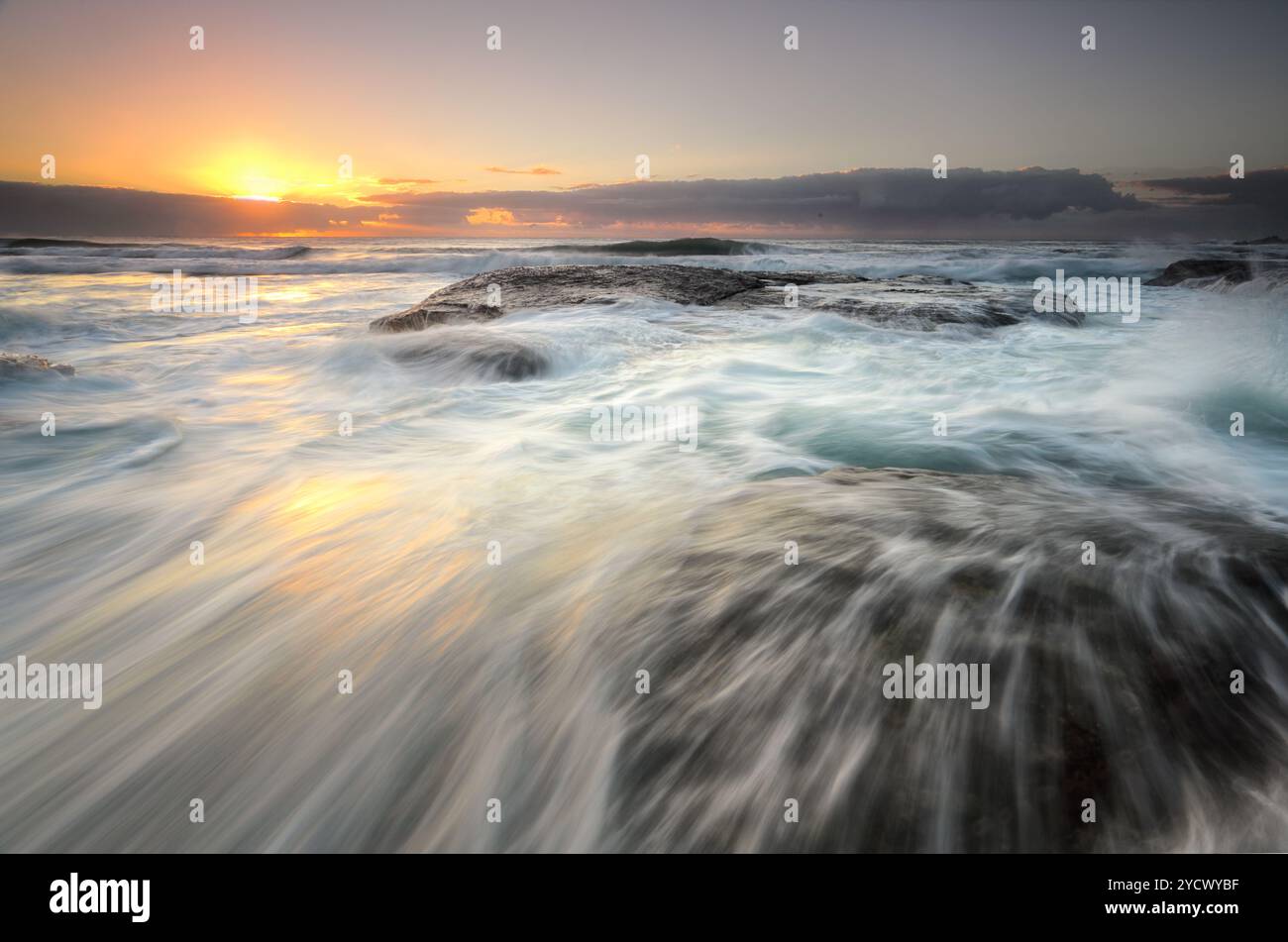 Ocean flowing rapidly over exposed rocks as the sun's glow peeks from ...