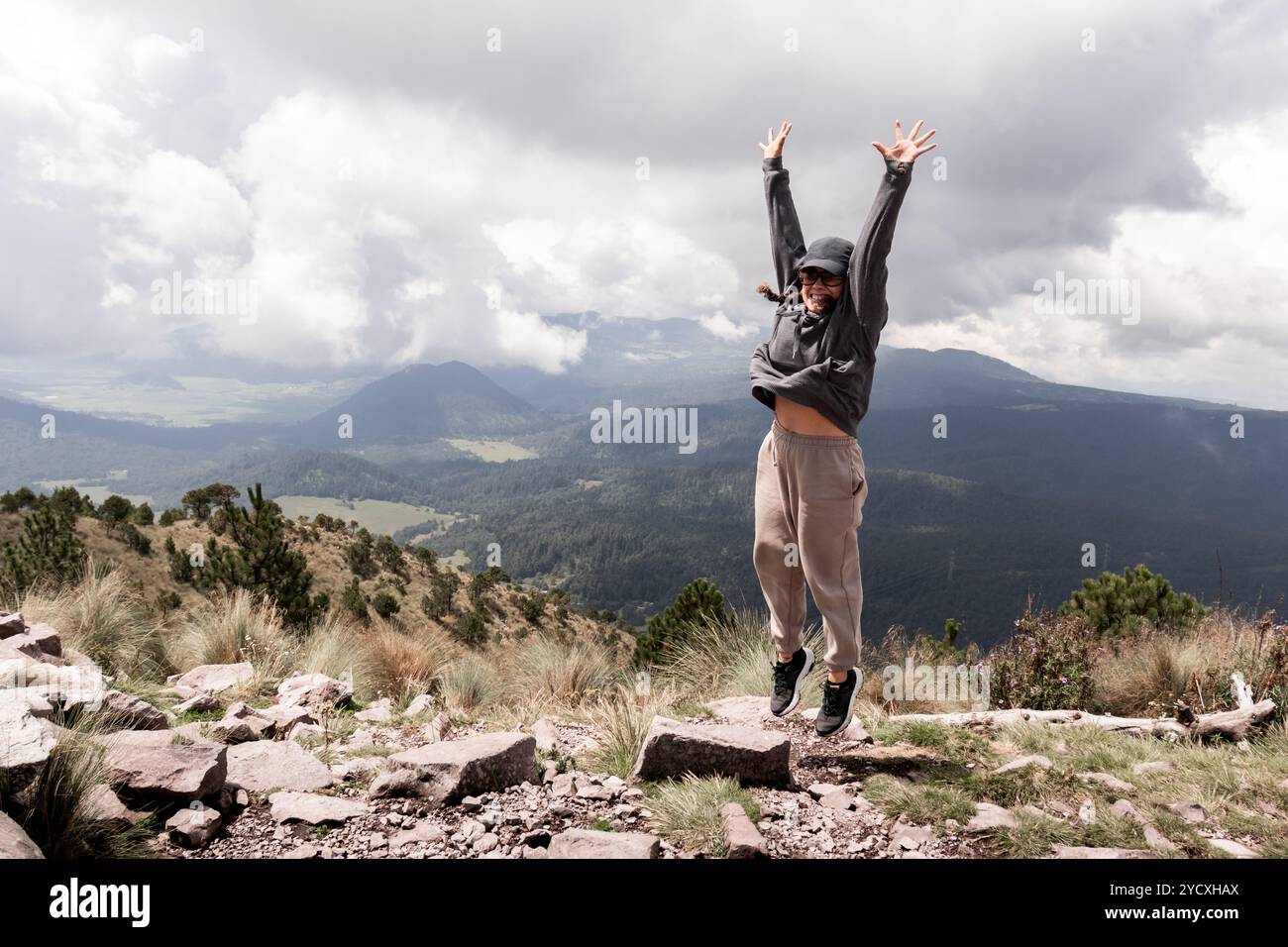 A happy young woman jumps joyfully, celebrating her hike at Cumbres del Ajusco, Mexico The lush landscape and dramatic clouds provide a stunning backd Stock Photo