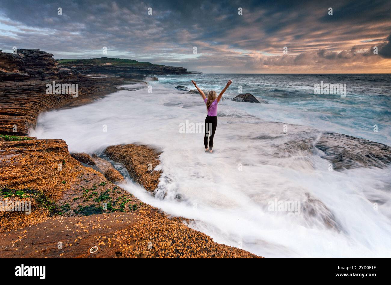 Girl standing on rocks as ocean waves wash past her feet Stock Photo