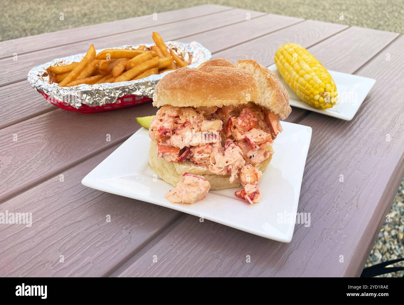 A portrait of a kaiser bun lobster roll with yellow corn and french fries. At Perry Long's Lobster Shack, Pound in Surry, Maine, USA. Stock Photo